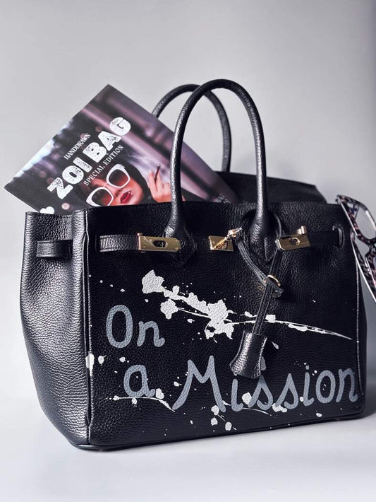 Designová černá kožená kabelka Zoi Bag s ručně psaným nápisem "On a Mission" a cákanci barvou. Zapínání zámkem turn-lock.