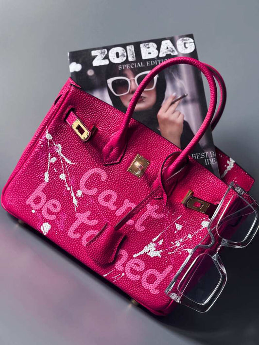 Designová růžová kožená kabelka Zoi Bag s ručně psaným nápisem a s cákanci barvou. Ikonické zapínání pomocí zámku s klíčkem.