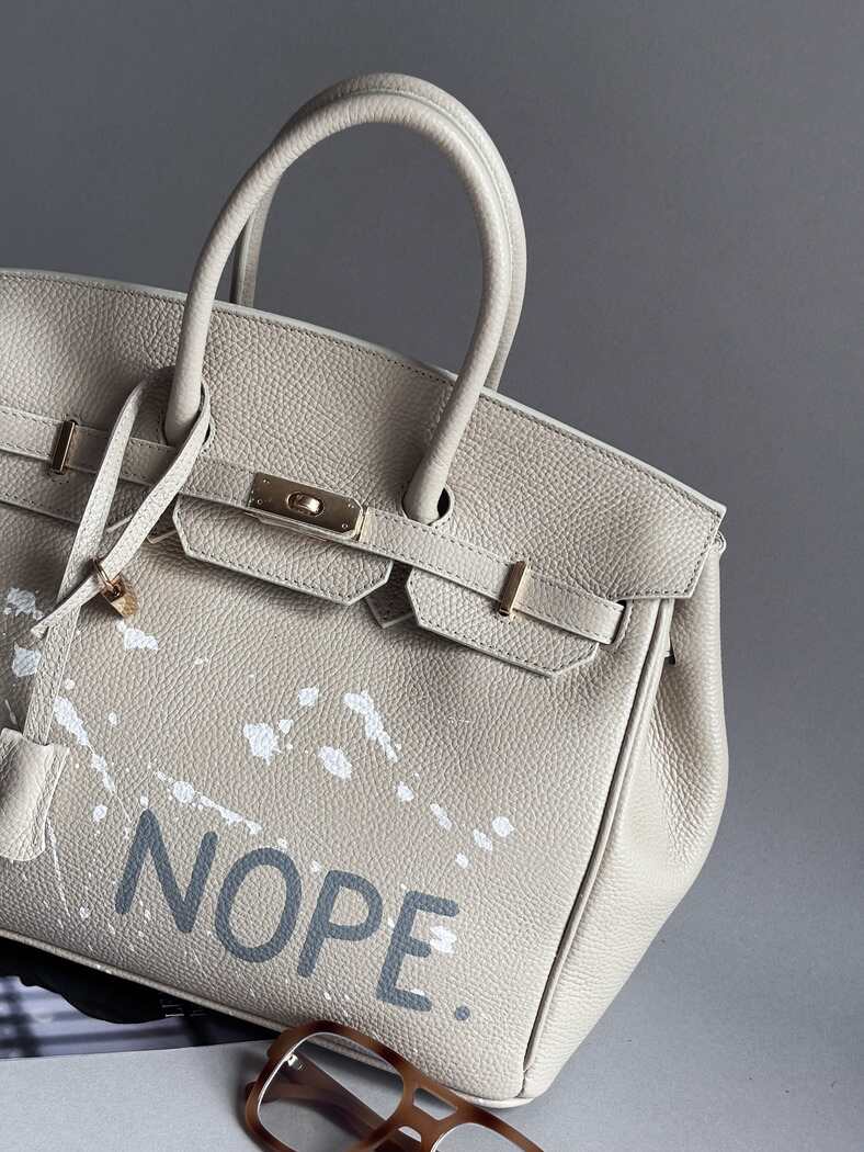 Béžová kožená kabelka Zoibag s ručně malovaným nápisem “Nope” a cákanci barvou. Zapnutá ikonickým zapínáním na zámek s klíčkem.