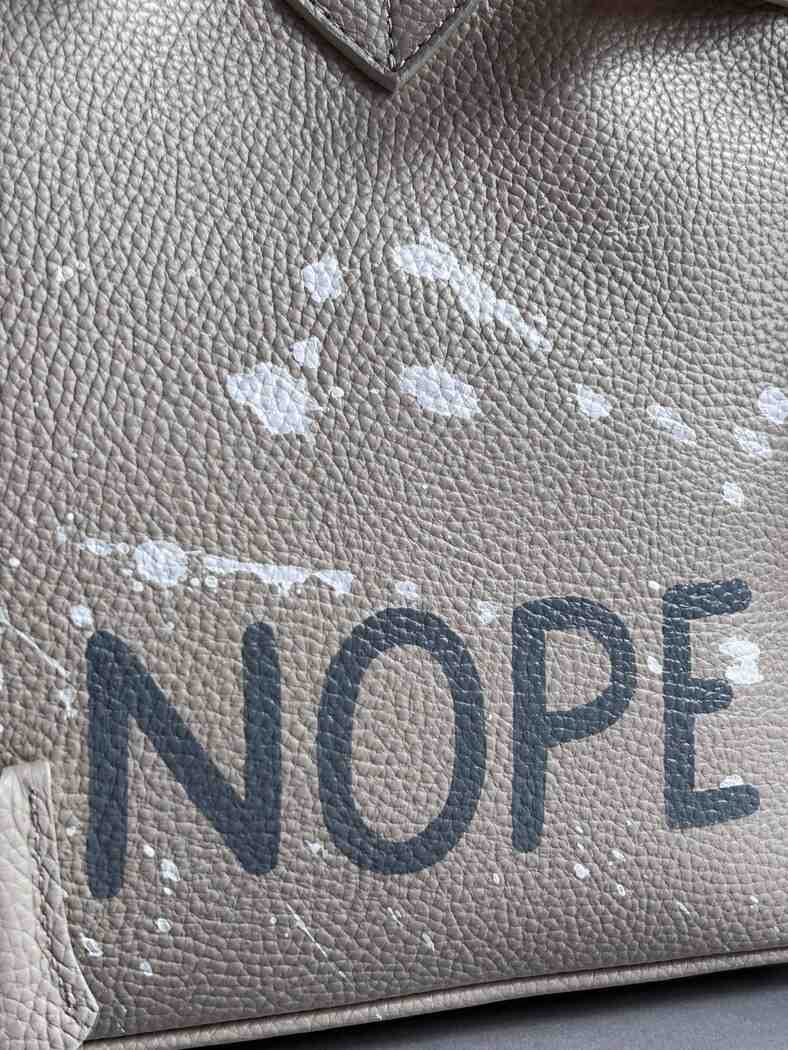 Béžová kožená kabelka Zoibag s ručně malovaným nápisem “Nope” a cákanci barvou. Zapnutá ikonickým zapínáním na zámek s klíčkem. Detail nápisu.