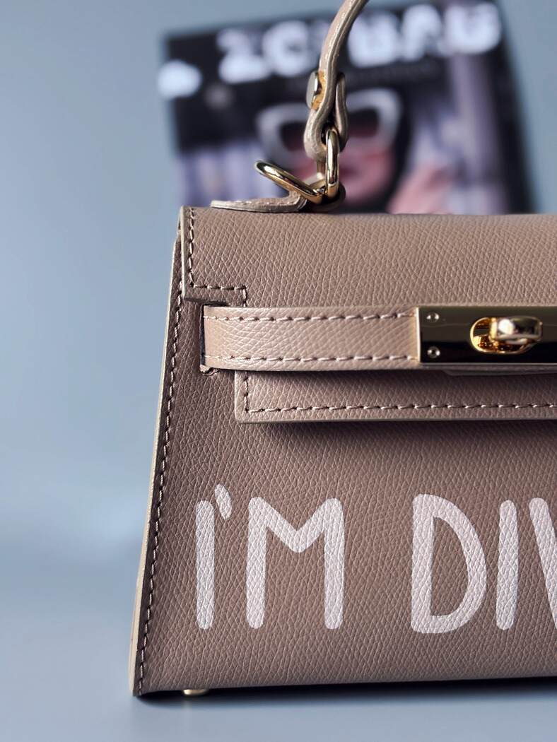 Designová luxusní béžová kabelka s ručně psaným nápisem, elegantním otočným zámkem a odnímatelným crossbody popruhem. Detail nápisu.
