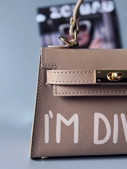 Designová luxusní béžová kabelka s ručně psaným nápisem, elegantním otočným zámkem a odnímatelným crossbody popruhem. Detail nápisu.