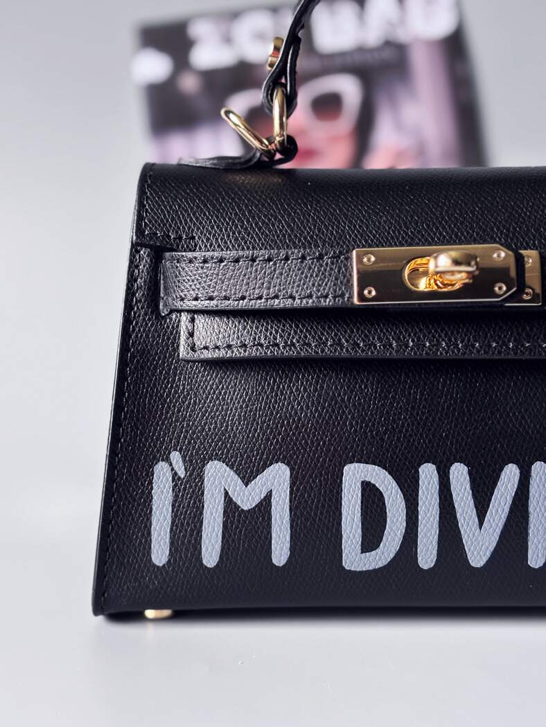 Designová luxusní černá kabelka s ručně psaným nápisem, elegantním otočným zámkem a odnímatelným crossbody popruhem. Detail nápisu.