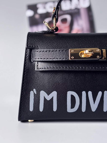 Designová luxusní černá kabelka s ručně psaným nápisem, elegantním otočným zámkem a odnímatelným crossbody popruhem. Detail nápisu.