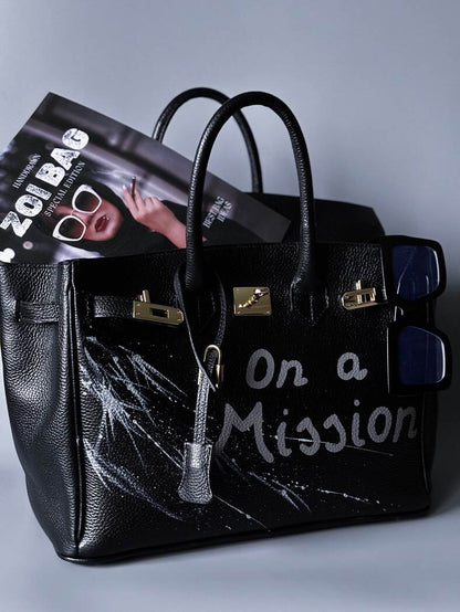 Černá kožená kabelka Zoi Bag s ručně psaným nápisem "On a Mision"  boku