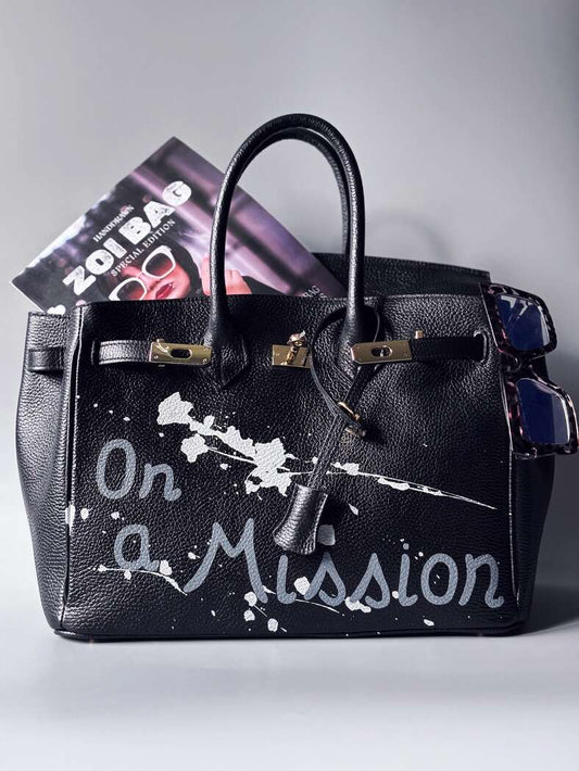Designová černá kožená kabelka Zoi Bag s ručně psaným nápisem "On a Mission" a cákanci barvou. Zapínání zámkem turn-lock.