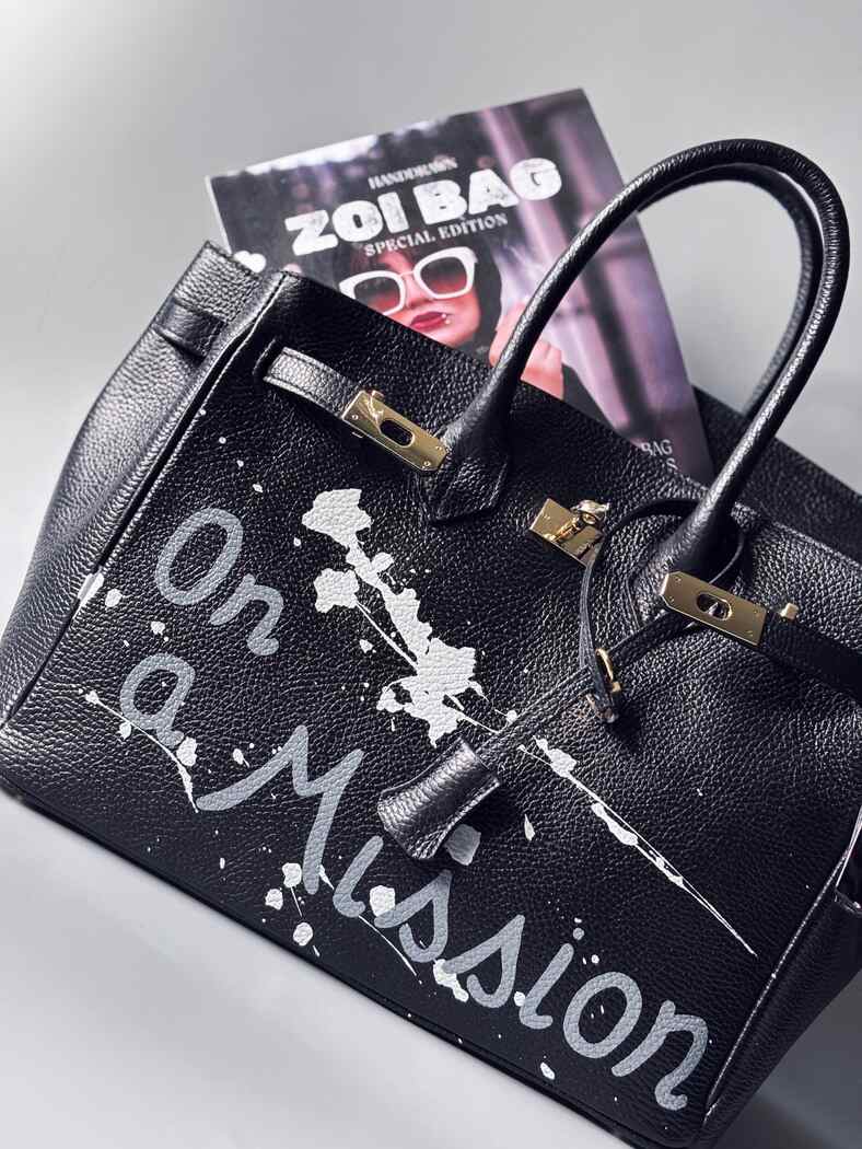 Designová černá kožená kabelka Zoi Bag s ručně psaným nápisem "On a Mission" a cákanci barvou. Zapínání zámkem turn-lock.