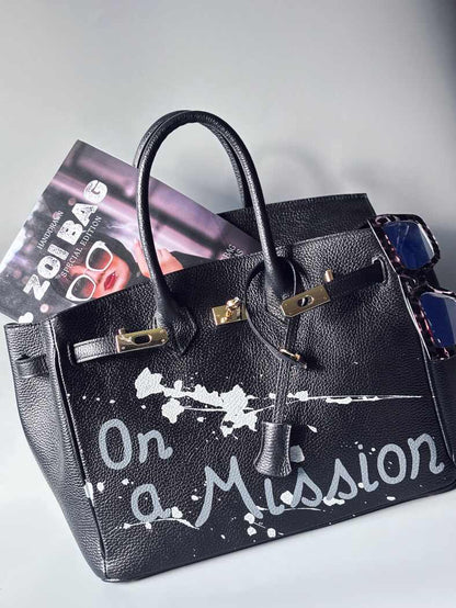 Designová černá kožená kabelka Zoi Bag s ručně psaným nápisem "On a Mission" a cákanci barvou. Zapínání zámkem turn-lock.