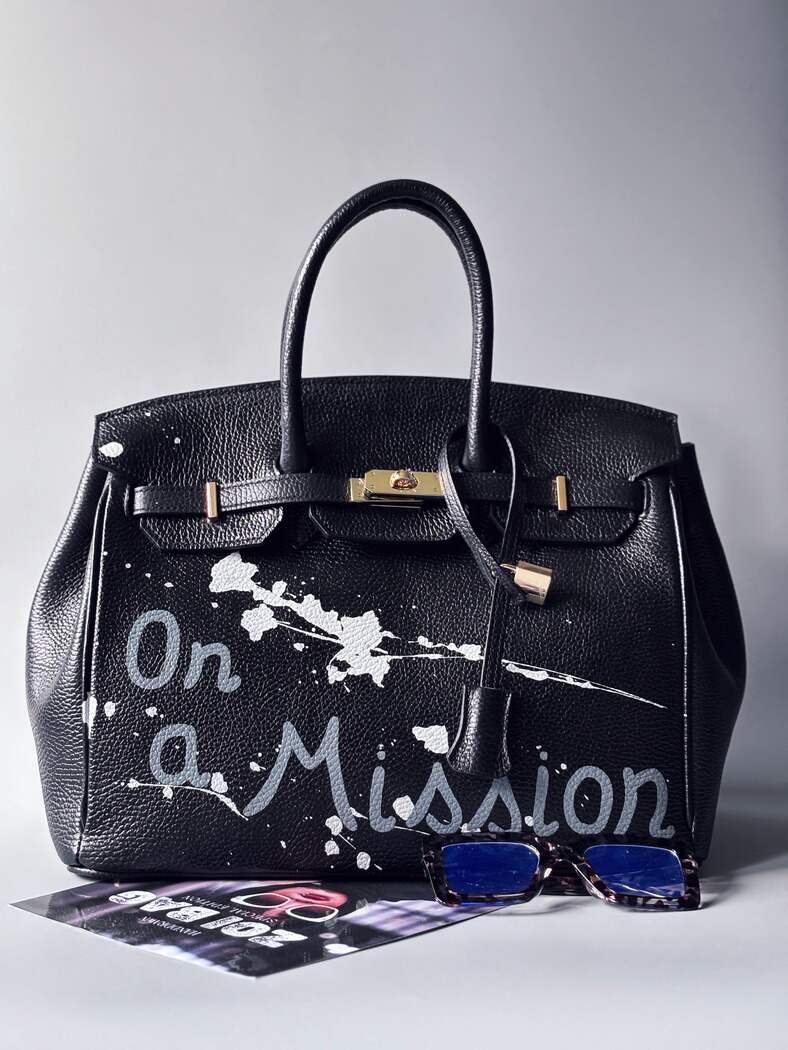 Designová černá kožená kabelka Zoi Bag s ručně psaným nápisem "On a Mission" a cákanci barvou. Zapínání zámkem turn-lock. Zapnutá.