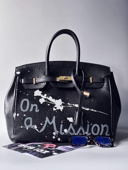 Designová černá kožená kabelka Zoi Bag s ručně psaným nápisem "On a Mission" a cákanci barvou. Zapínání zámkem turn-lock. Zapnutá.