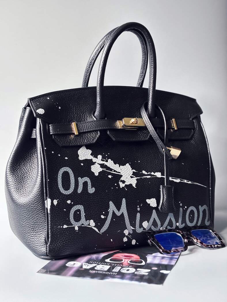 Designová černá kožená kabelka Zoi Bag s ručně psaným nápisem "On a Mission" a cákanci barvou. Zapínání zámkem turn-lock. Zapnutá.