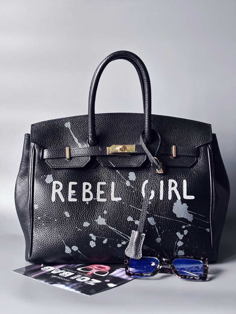 Designová černá kožená kabelka Zoi Bag s ručně psaným nápisem "Rebel Girl" a cákanci barvou. Zámek s klíčkem na zapnutí kabelky.