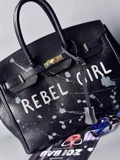 Designová černá kožená kabelka Zoi Bag s ručně psaným nápisem "Rebel Girl" a cákanci barvou. Zámek s klíčkem na zapnutí kabelky. Zapnutá.