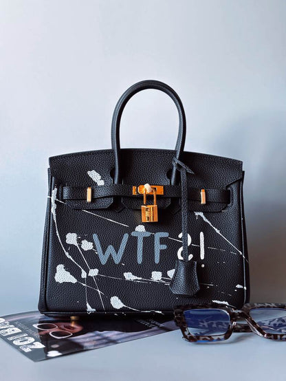 Designová černá kožená kabelka Zoi Bag z právé kůže s ručně psaným nápisem a cákanci barvou. Zlatý zámek s klíčkem. Zapnutá.