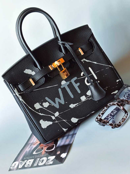 Designová černá kožená kabelka Zoi Bag z právé kůže s ručně psaným nápisem a cákanci barvou. Zlatý zámek s klíčkem. Zapnutá.