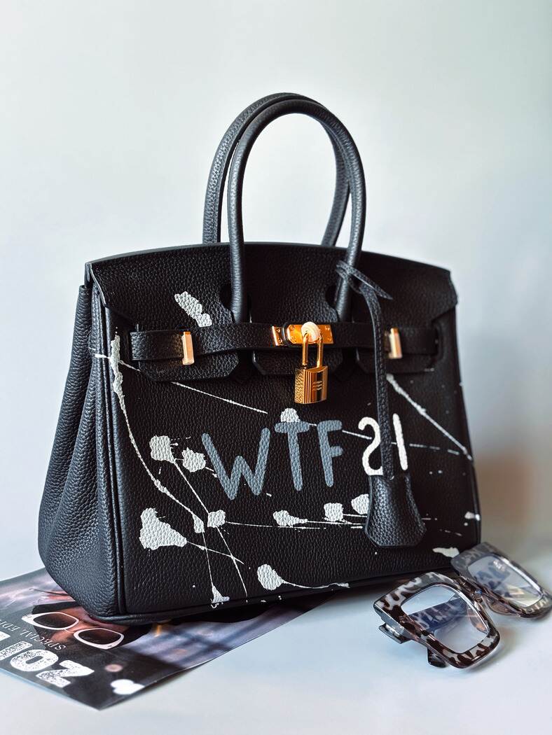 Designová černá kožená kabelka Zoi Bag z právé kůže s ručně psaným nápisem a cákanci barvou. Zlatý zámek s klíčkem. Zapnutá.