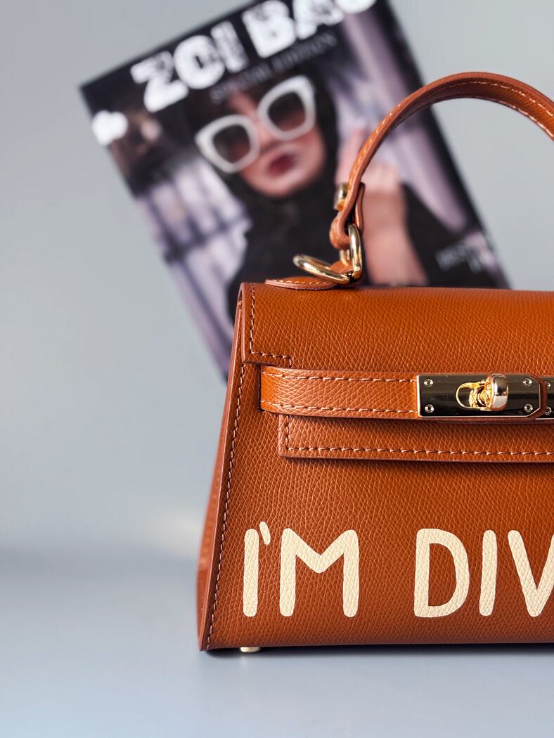 Designová luxusní hnědá kabelka s ručně psaným nápisem, elegantním otočným zámkem a odnímatelným crossbody popruhem. Detail nápisu.