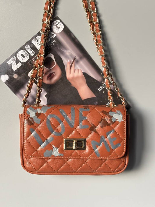 Hnědá kožená kabelka Zoi Bag s ručně psaným nápisem "Love Me" a s cákanci barvu.