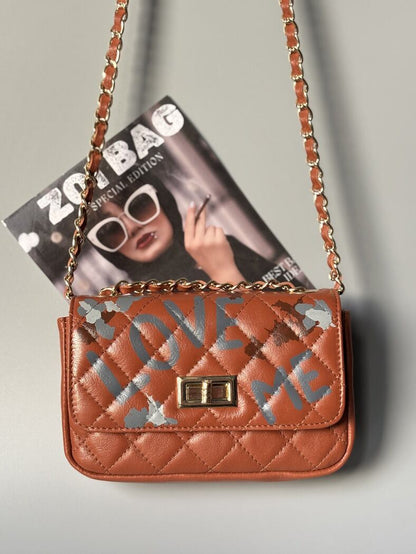 Hnědá kožená kabelka Zoi Bag s ručně psaným nápisem "Love Me" a s cákanci barvu.