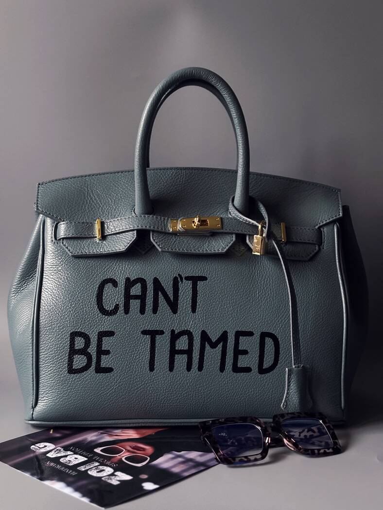 Kožená šedomodrá kabelka Zoi Bag s ručně psaným nápisem "Can't be tamed" zapnutá