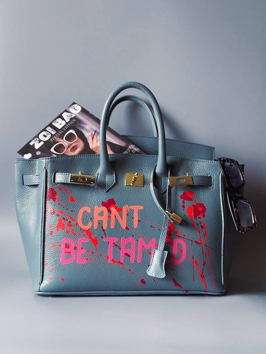 Modrá/šedá kožená kabelka Zoi Bag s ručně psaným nápisem "Can't be tamed" a barevnými cákanci. Doplněna zámkem s klíčkem.