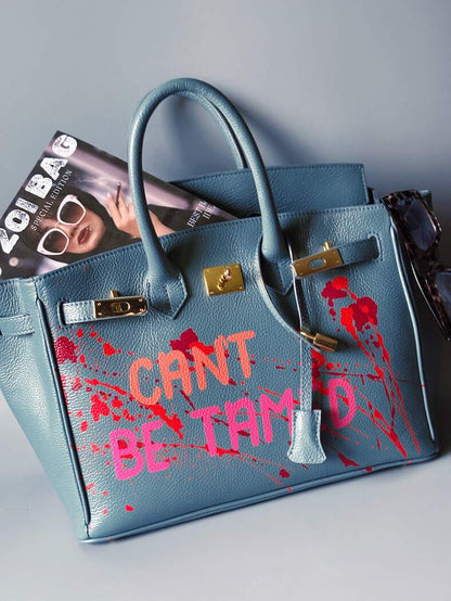 Modrá/šedá kožená kabelka Zoi Bag s ručně psaným nápisem "Can't be tamed" a barevnými cákanci. Doplněna zámkem s klíčkem.