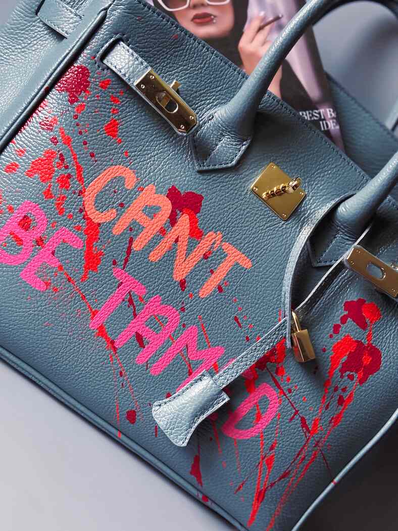 Modrá/šedá kožená kabelka Zoi Bag s ručně psaným nápisem "Can't be tamed" a barevnými cákanci. Doplněna zámkem s klíčkem.