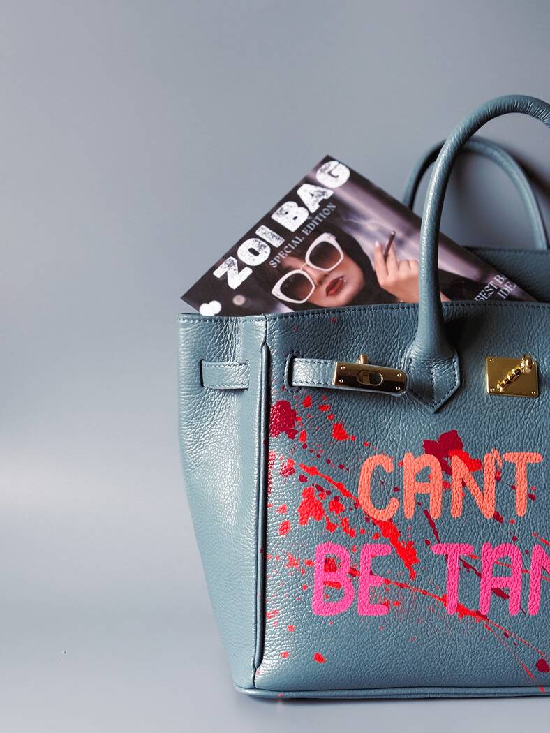 Modrá/šedá kožená kabelka Zoi Bag s ručně psaným nápisem "Can't be tamed" a barevnými cákanci. Doplněna zámkem s klíčkem.