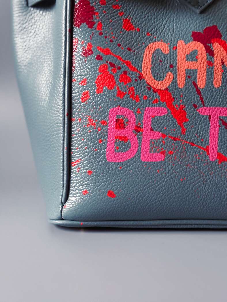 Modrá/šedá kožená kabelka Zoi Bag s ručně psaným nápisem "Can't be tamed" a barevnými cákanci. Doplněna zámkem s klíčkem. Detail nápisu