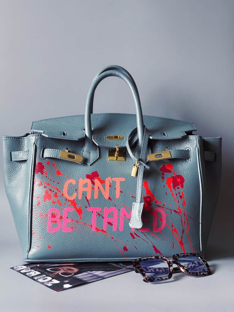 Modrá/šedá kožená kabelka Zoi Bag s ručně psaným nápisem "Can't be tamed" a barevnými cákanci. Doplněna zámkem s klíčkem.