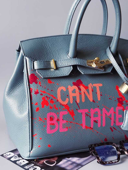 Modrá/šedá kožená kabelka Zoi Bag s ručně psaným nápisem "Can't be tamed" a barevnými cákanci. Doplněna zámkem s klíčkem.