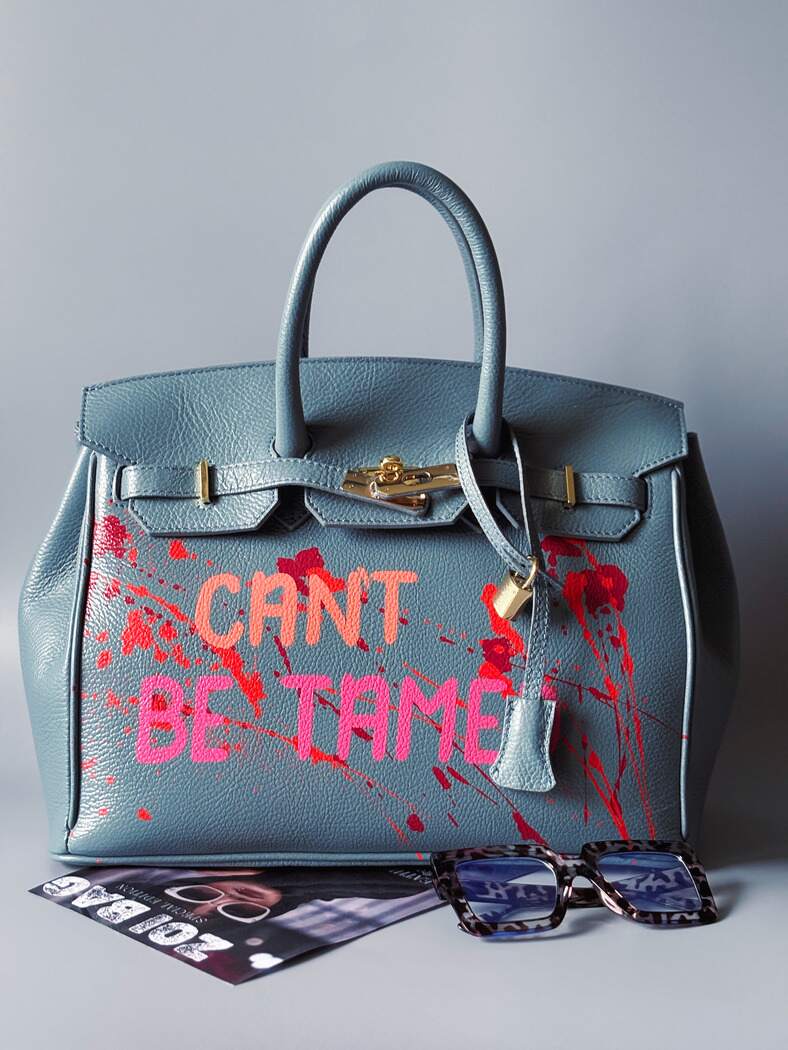 Modrá/šedá kožená kabelka Zoi Bag s ručně psaným nápisem "Can't be tamed" a barevnými cákanci. Doplněna zámkem s klíčkem.