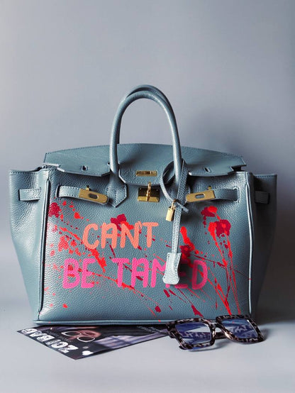 Modrá/šedá kožená kabelka Zoi Bag s ručně psaným nápisem "Can't be tamed" a barevnými cákanci. Doplněna zámkem s klíčkem.