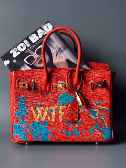 Oranžová kožená kabelka Zoi Bag s ručně psaným nápisem "WTF" a modrými cákanci barvou. Zapínání pomocí zlatého zámku s klíčkem.