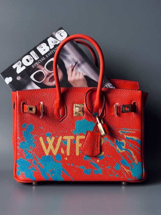 Oranžová kožená kabelka Zoi Bag s ručně psaným nápisem "WTF" a modrými cákanci barvou. Zapínání pomocí zlatého zámku s klíčkem.