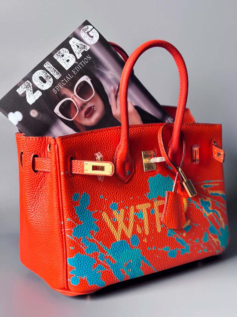 Oranžová kožená kabelka Zoi Bag s ručně psaným nápisem "WTF" a modrými cákanci barvou. Zapínání pomocí zlatého zámku s klíčkem.