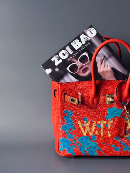 Designová oranžová kožená kabelka Zoi Bag s ručně psaným nápisem "WTF" a modrými cákanci barvou. Zapínání pomocí zlatého zámku s klíčkem.
