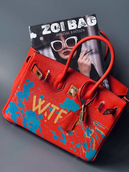Oranžová kožená kabelka Zoi Bag s ručně psaným nápisem "WTF" a modrými cákanci barvou. Zapínání pomocí zlatého zámku s klíčkem.