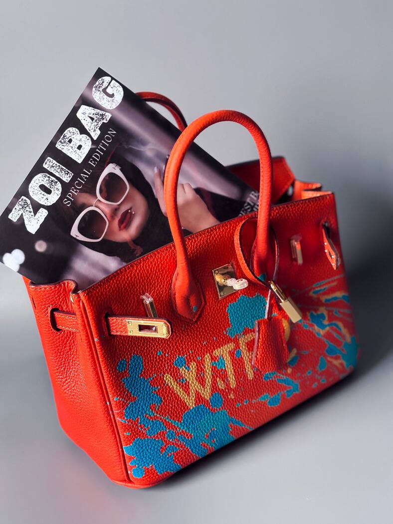 Oranžová kožená kabelka Zoi Bag s ručně psaným nápisem "WTF" a modrými cákanci barvou. Zapínání pomocí zlatého zámku s klíčkem.