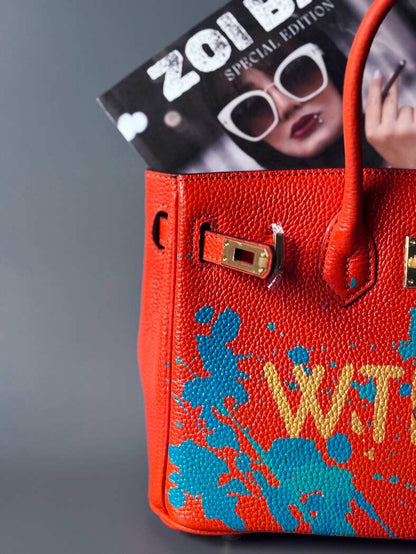 Oranžová kožená kabelka Zoi Bag s ručně psaným nápisem "WTF" a modrými cákanci barvou. Zapínání pomocí zlatého zámku s klíčkem. Detail.