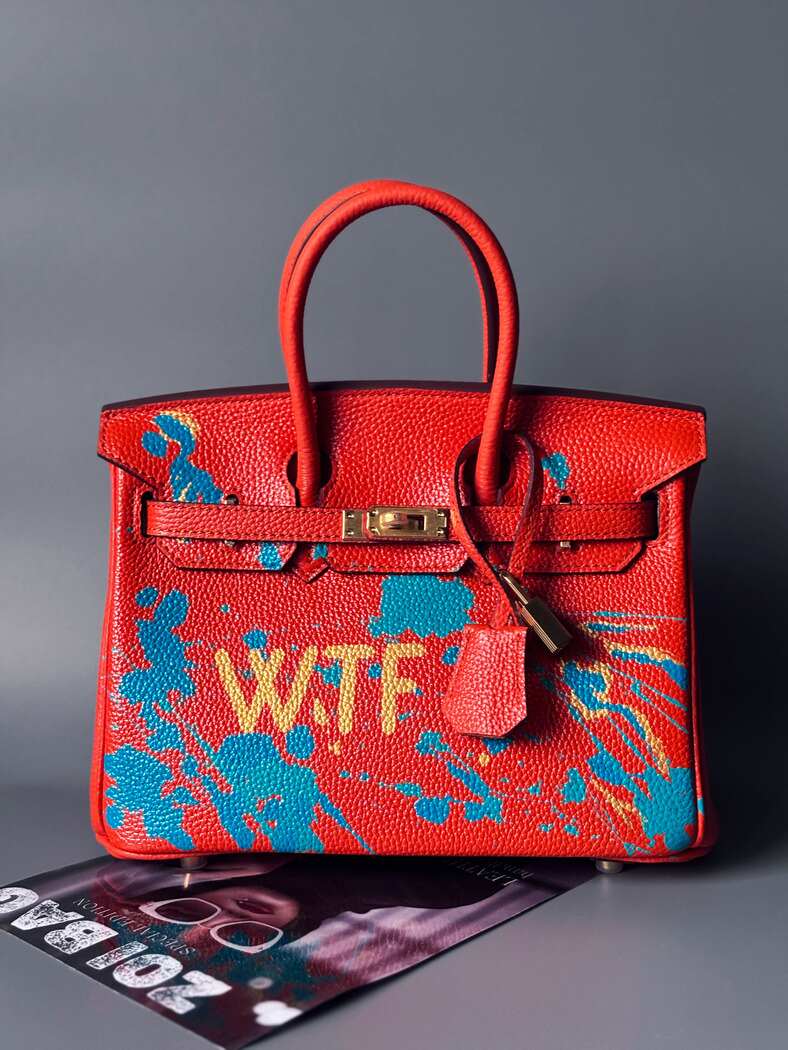 Oranžová kožená kabelka Zoi Bag s ručně psaným nápisem "WTF" a modrými cákanci barvou. Zapínání pomocí zlatého zámku s klíčkem. Zapnutá.