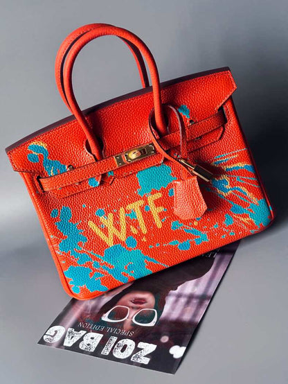 Oranžová kožená kabelka Zoi Bag s ručně psaným nápisem "WTF" a modrými cákanci barvou. Zapínání pomocí zlatého zámku s klíčkem. Zapnutá.