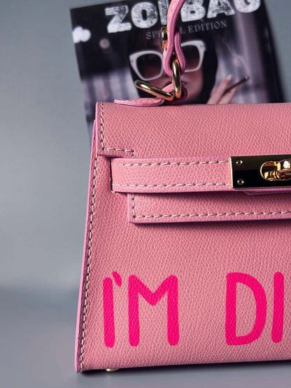 Designová luxusní růžová kabelka s ručně psaným nápisem, elegantním otočným zámkem a odnímatelným crossbody popruhem. Detail nápisu.