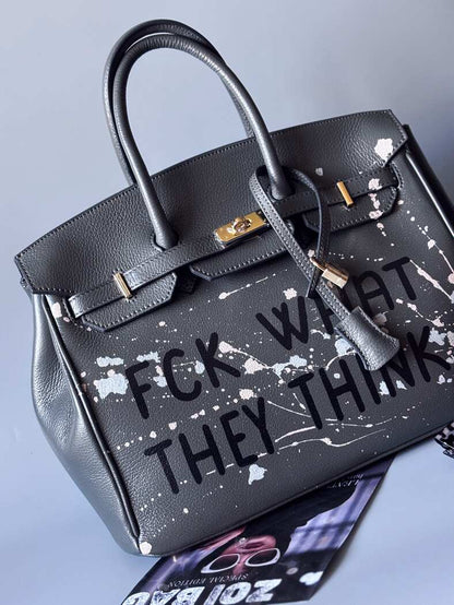 Designová luxusní šedá kožená kabelka Zoi Bag s ručně psaným nápisem a s cákanci barvou. Zapínání pomocí klíčku a zámku. Zapnutá.