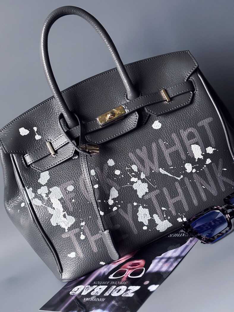 Tmavě šedá kožená kabelka Zoi Bag s ručně psaným nápisem "FCK what they think" a cákanci barvou. Zapnutá.