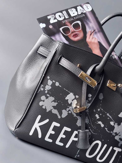 Luxusní designová kožená kabelka Zoi Bag s ručně psaným nápisem "Keep Out" a cákanci barvou. Zapínání pomocí zámku s klíčkem.